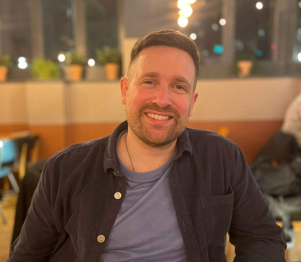 Image of Daniel, a white, bearded man in his early 40s, smiling into the camera in a relaxed setting.